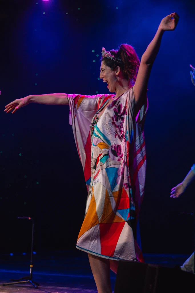 Daphne Always is smiling and joyfully dancing on stage with her arms raised, wearing a colorful, patterned dress and a floral crown in her hair, at JoCo Cruise 2025.