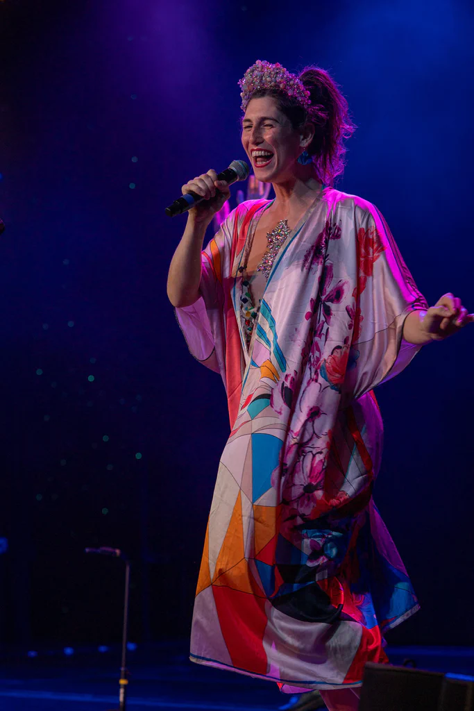 Daphne Always, at JoCo Cruise 2025, is performing on stage, smiling and holding a microphone, wearing a colorful, patterned dress with floral and geometric designs, and a floral headpiece with bright pink accents.