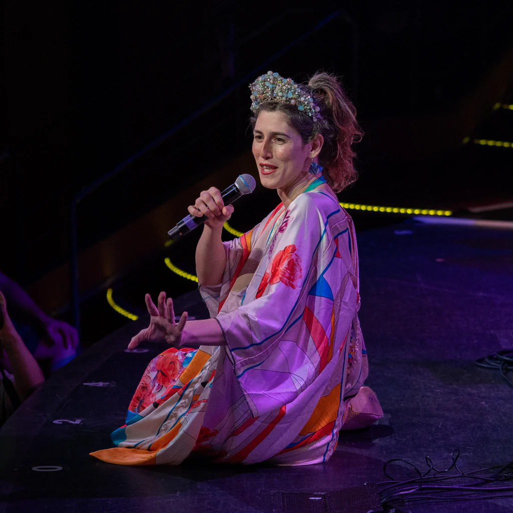 Daphne Always, wearing a colorful kimono and a decorative headpiece, sits on stage holding a microphone, engaging with the audience during JoCo Cruise 2025.