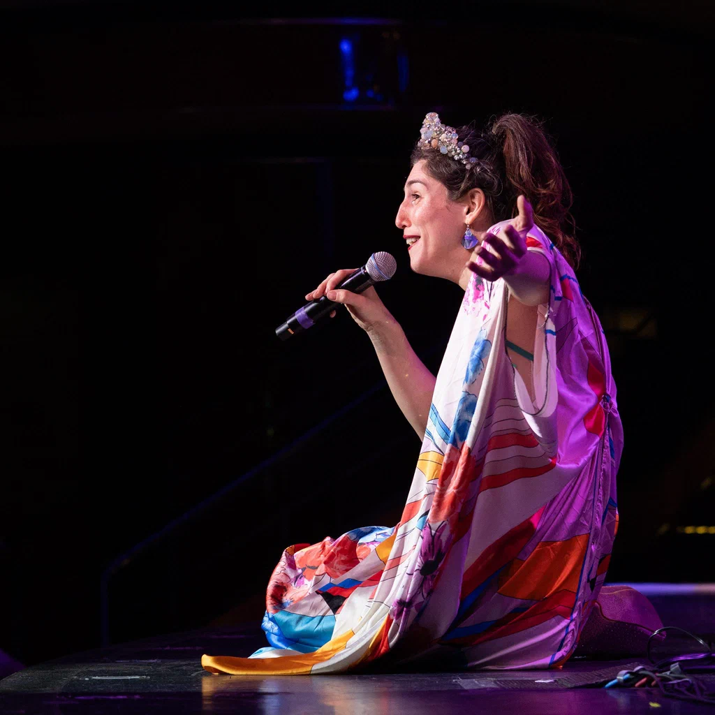 Daphne Always, wearing a colorful, patterned outfit and a sparkly headband, sits on stage at JoCo Cruise 2025, holding a microphone and gesturing with her hand.