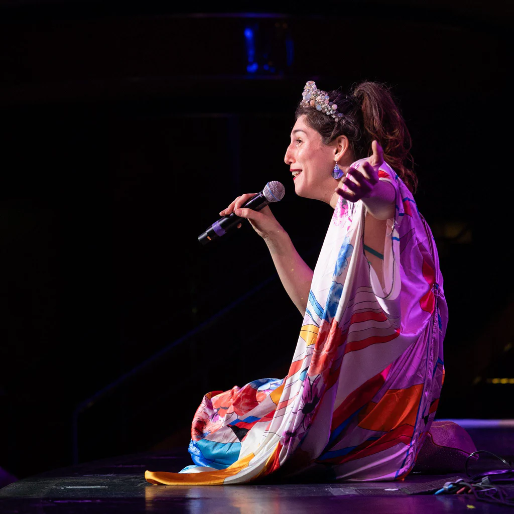 Daphne Always, wearing a colorful, patterned outfit and a sparkly headband, sits on stage at JoCo Cruise 2025, holding a microphone and gesturing with her hand.