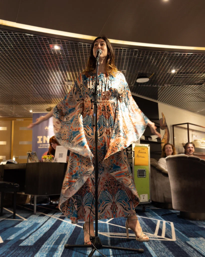 Daphne Always performing on stage with outstretched arms, wearing a flowing, patterned dress and high heels, at JoCo Cruise 2025.