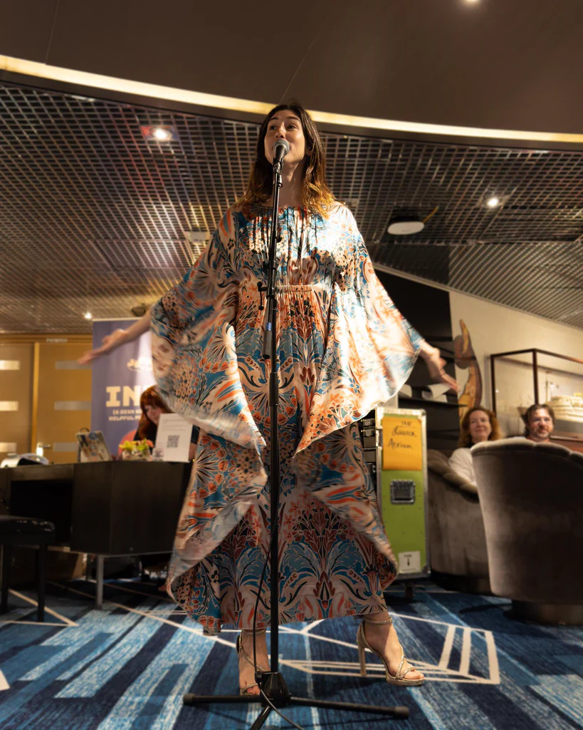 Daphne Always performing on stage with outstretched arms, wearing a flowing, patterned dress and high heels, at JoCo Cruise 2025.