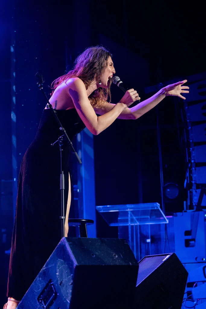 Daphne Always performing passionately on stage at JoCo Cruise 2025, wearing a black dress and singing into a microphone.