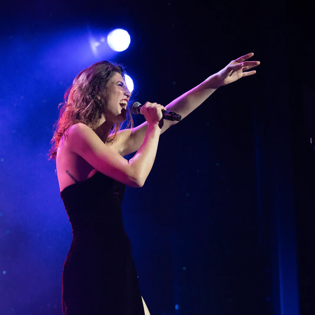 Daphne Always performs on stage at JoCo Cruise 2025, singing into a microphone with her right arm outstretched and a joyful expression.