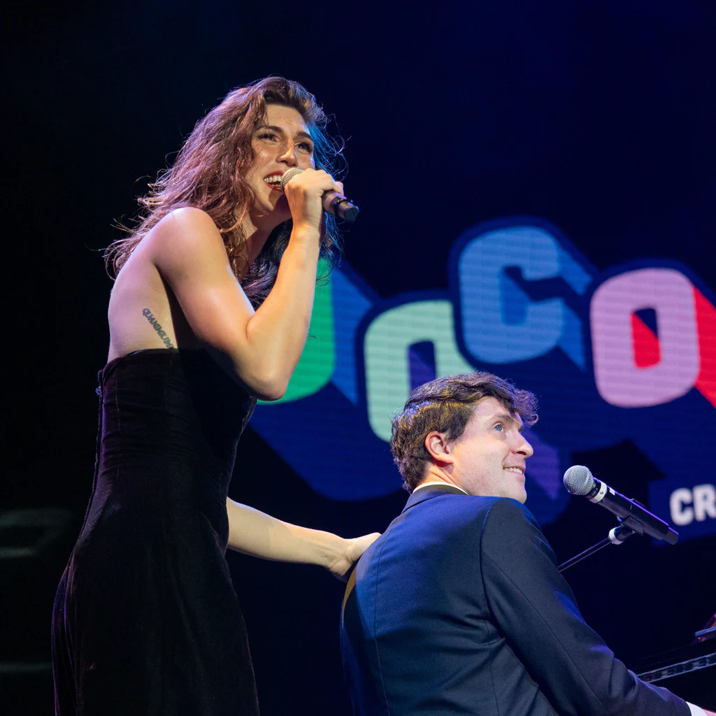 A woman with curly hair, labeled Daphne Always, performs on stage with a microphone, standing beside a man at a piano with a colorful "JoCo" logo in the background.