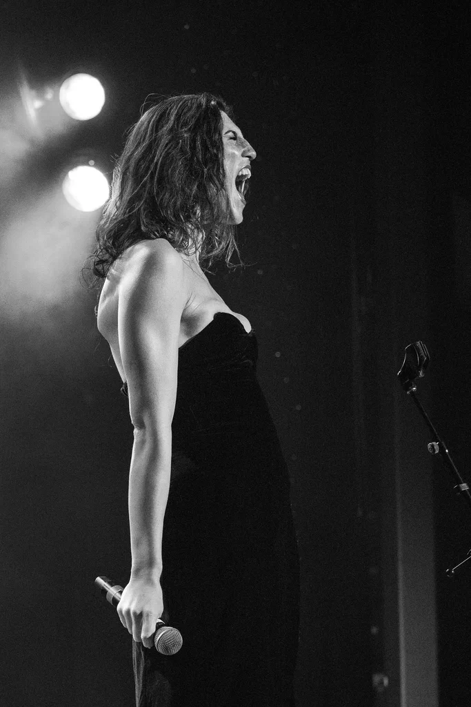 A woman, identified as Daphne Always, passionately performs on stage at JoCo Cruise 2025, holding a microphone in her right hand, with her mouth open and hair tousled.