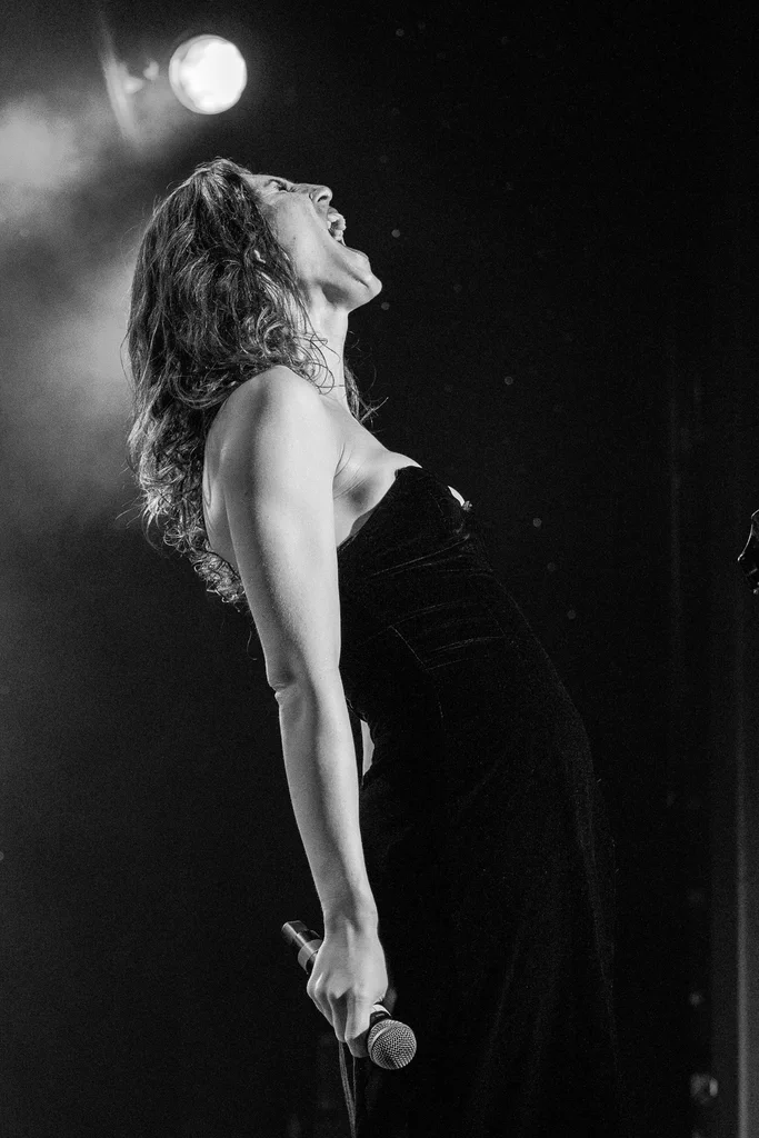 Daphne Always, on JoCo Cruise 2025, is performing passionately on stage, holding a microphone in her right hand, with her head tilted back and mouth open in song or expression.