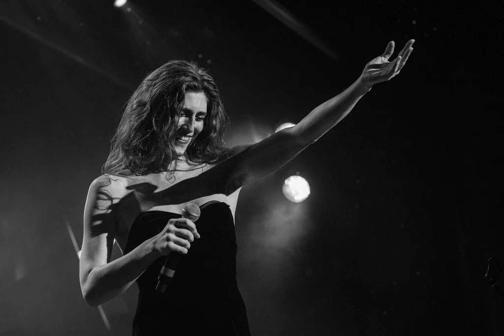 A woman, identified as Daphne Always, smiling and holding a microphone on stage during JoCo Cruise 2025, with her arm raised in the air.