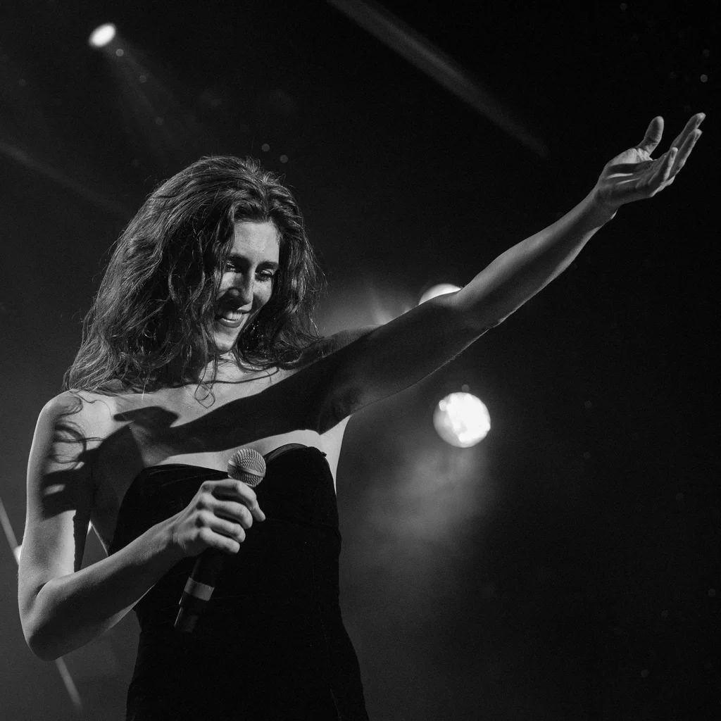 A woman named Daphne Always, with wavy hair, is smiling on stage while holding a microphone, reaching out with her arm extended.