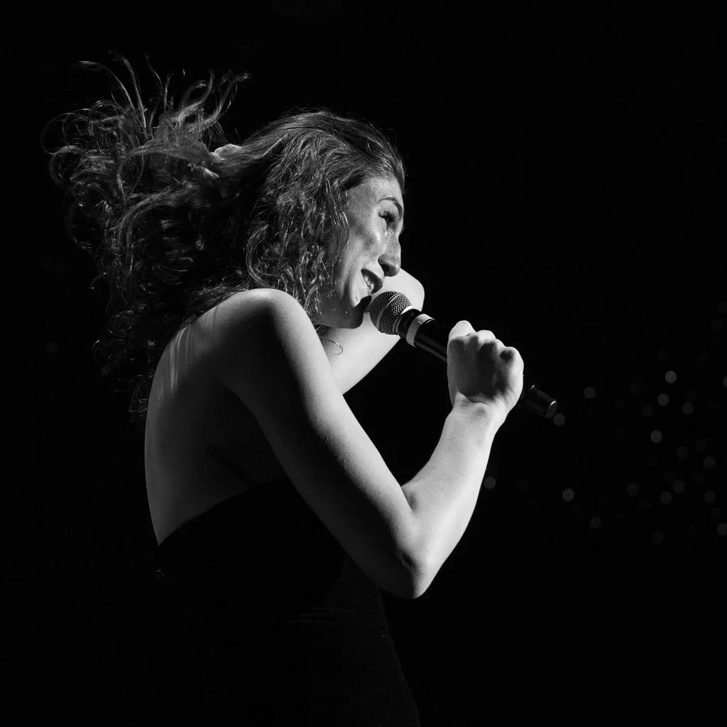 Daphne Always singing into a microphone on JoCo Cruise 2025, with her curly hair flowing and expressive face.