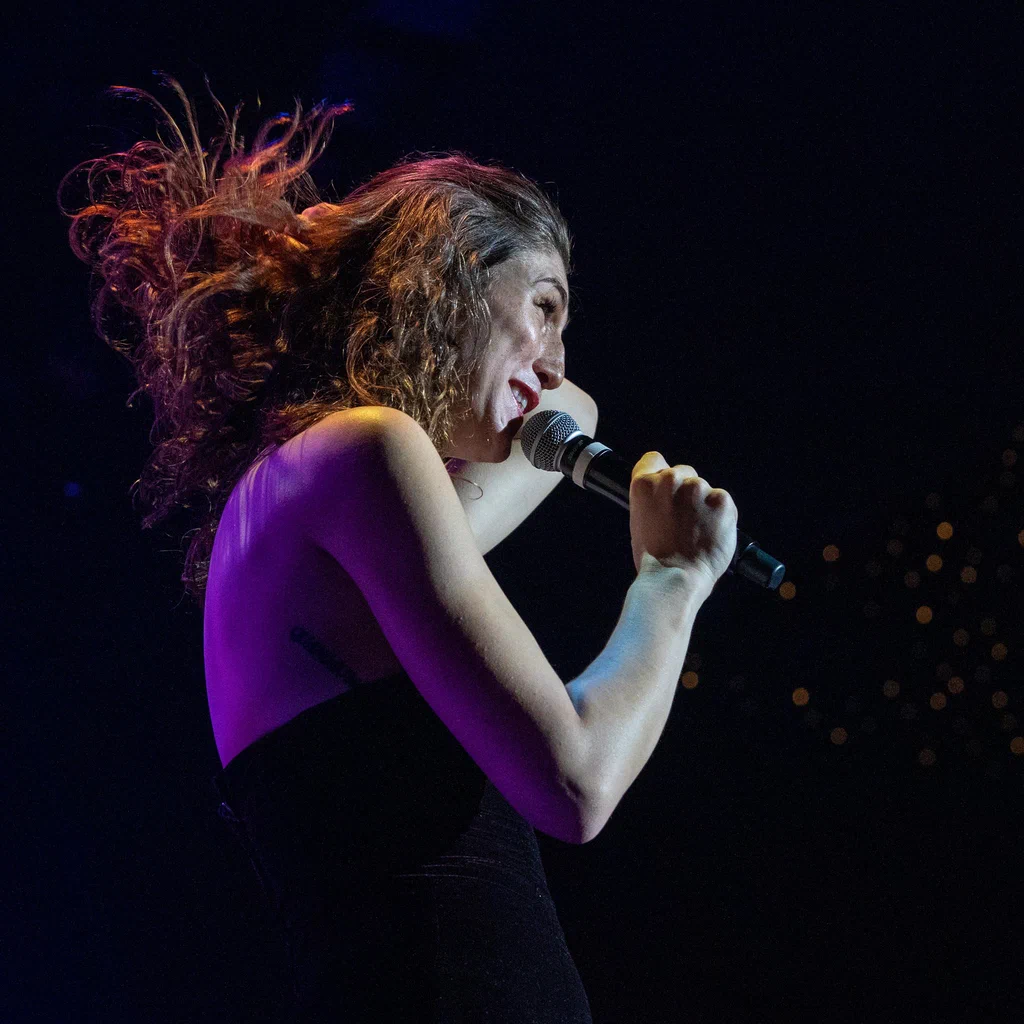 A woman with curly hair, named Daphne Always, singing into a microphone on JoCo Cruise 2025 stage, with colorful lighting and dark background.