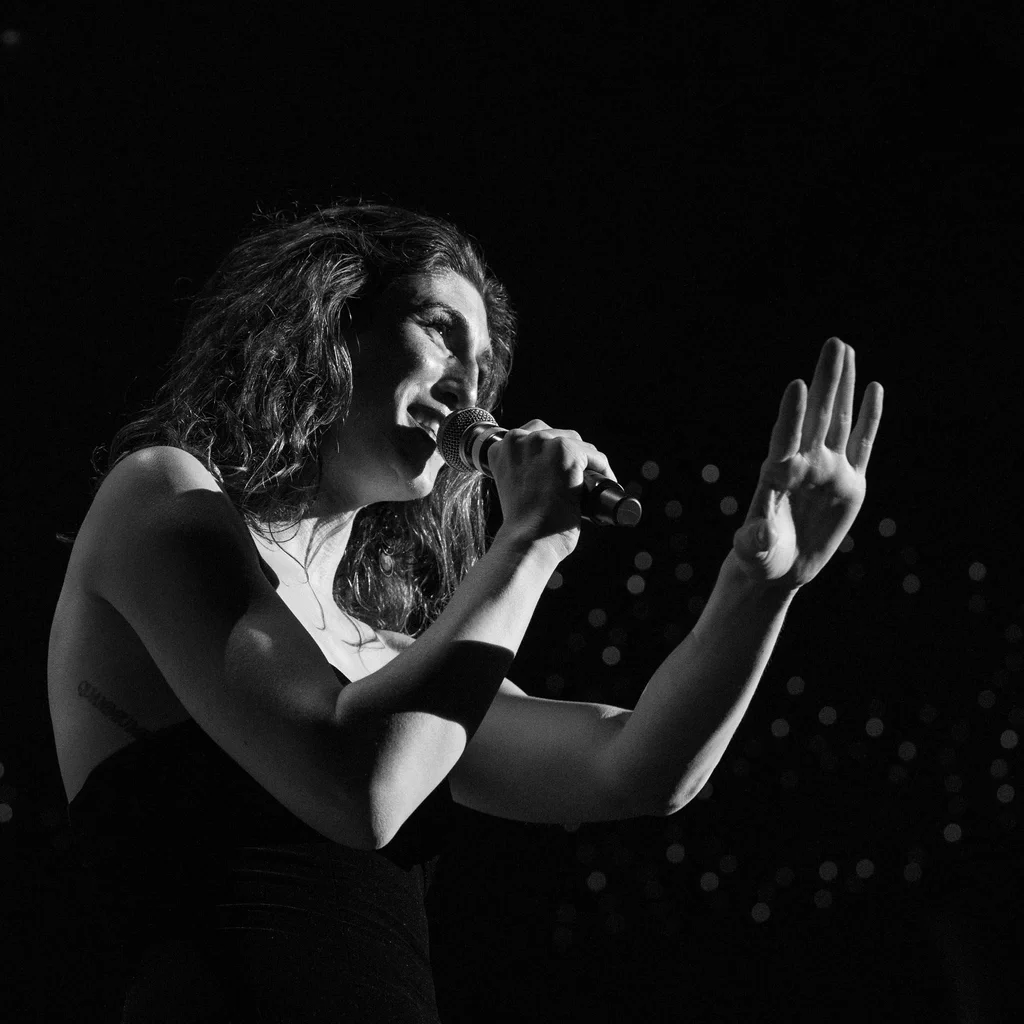 A woman, identified as Daphne Always, singing into a microphone on the JoCo Cruise 2025, with her hand raised in a gesture.