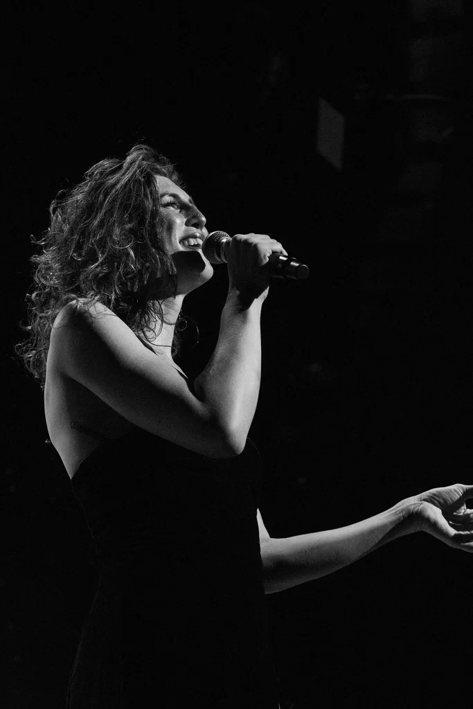 A woman, named Daphne Always, performs on stage at JoCo Cruise 2025, singing into a microphone with a joyful expression.
