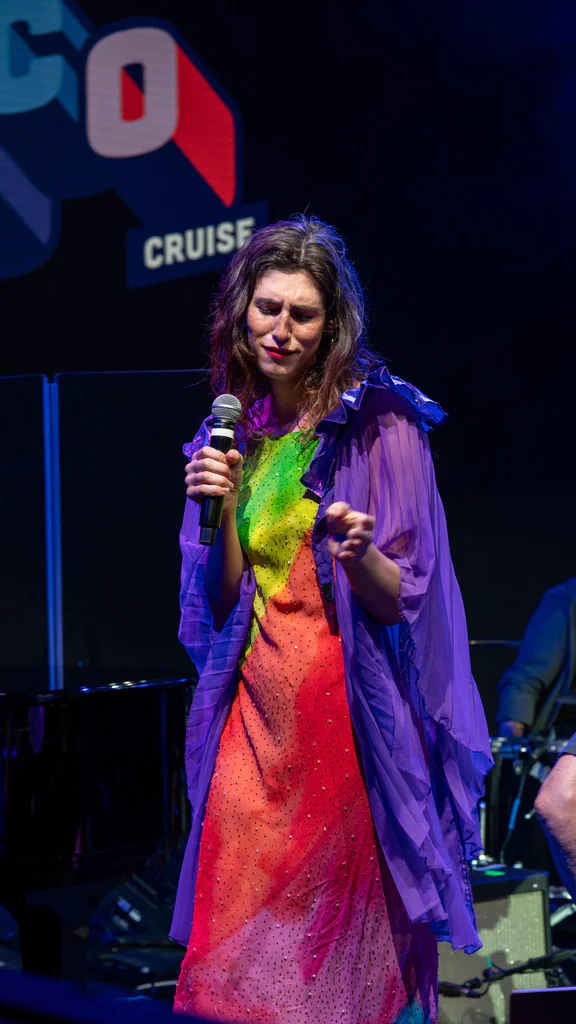 A woman named Daphne Always performing on stage at JoCo Cruise 2025, wearing a colorful dress and a purple sheer overlay, holding a microphone with a concentrated expression.