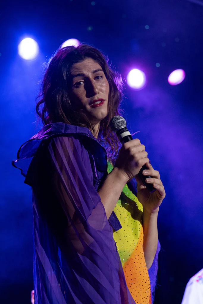Daphne Always performing on stage at JoCo Cruise 2025, holding a microphone with purple and colorful background lighting.