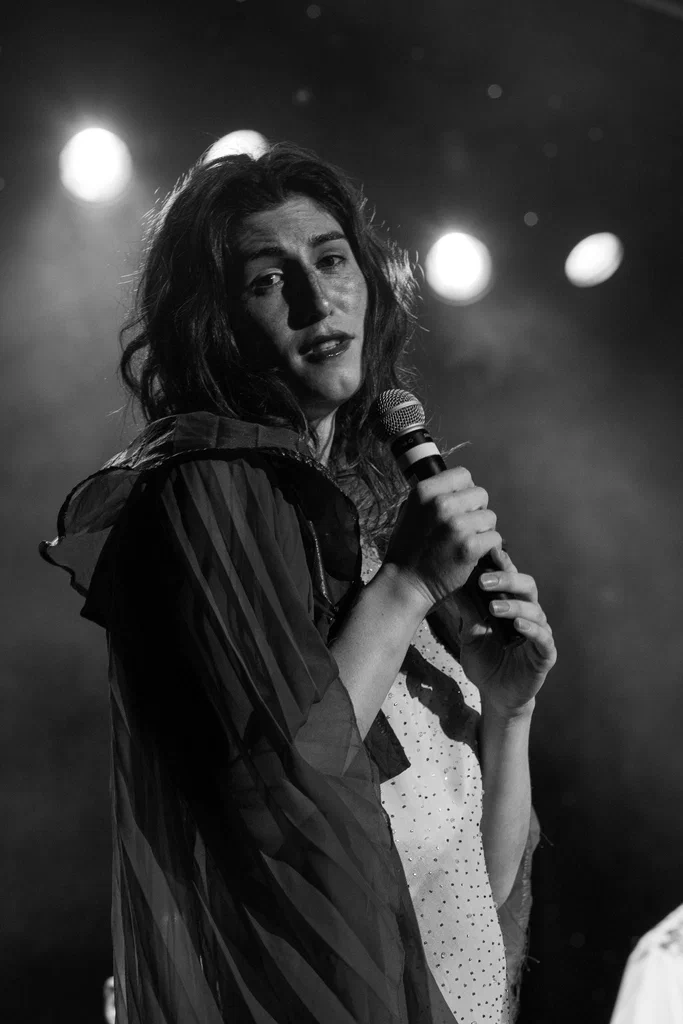 Daphne Always on JoCo Cruise 2025 performing or speaking on stage with a microphone, illuminated by stage lights in a black-and-white photo.