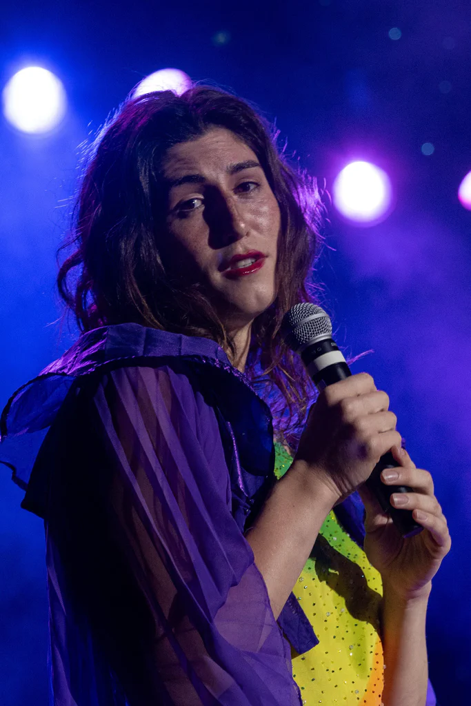 Daphne Always holding a microphone on stage during JoCo Cruise 2025, with colorful stage lights in the background.