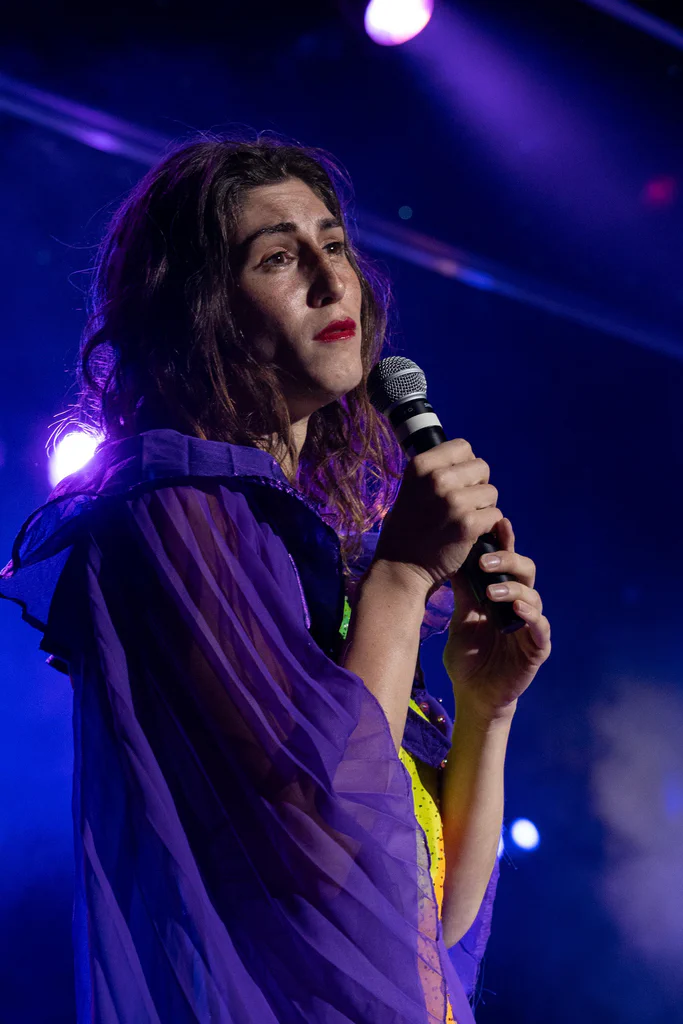 Daphne Always performing on stage during JoCo Cruise 2025, holding a microphone with purple stage lighting in the background.