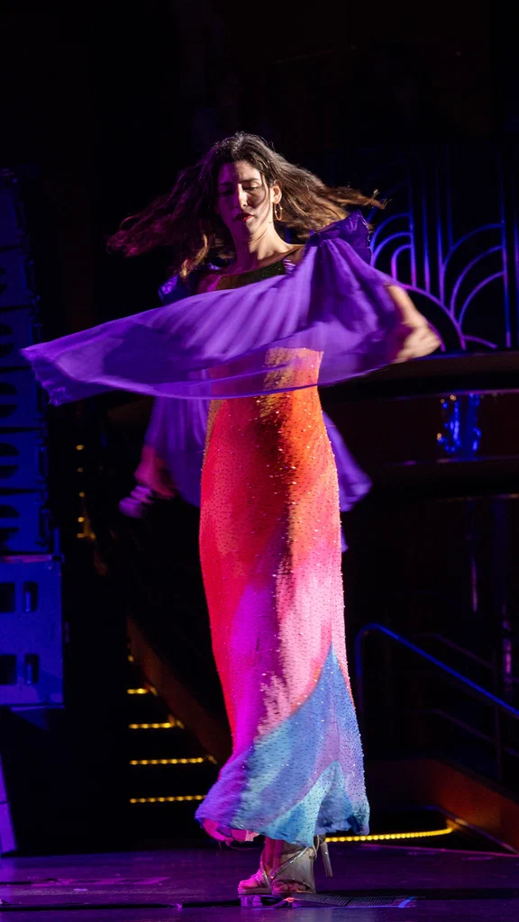 Daphne Always performs a dance on stage, wearing a fitted, colorful gown with sequins, under vibrant stage lighting.