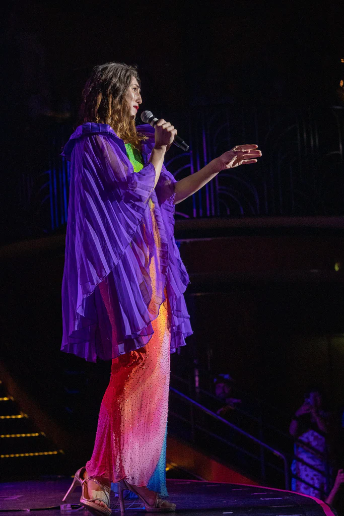 Daphne Always performing on stage at JoCo Cruise 2025, wearing a colorful outfit with a purple sheer cape and a multicolored dress, holding a microphone.