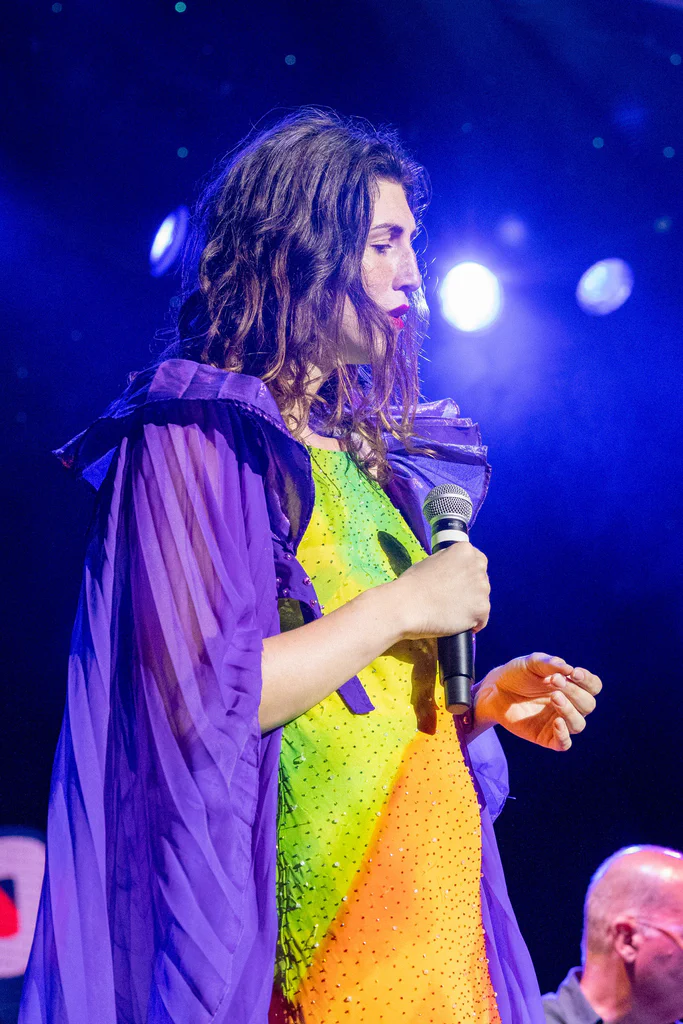 Daphne Always, a woman with wavy brown hair, is holding a microphone on stage during JoCo Cruise 2025, wearing a colorful, sparkly dress and a purple sheer cape.