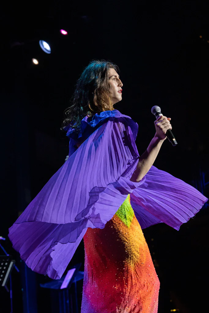 Daphne Always performing on stage at JoCo Cruise 2025, wearing a vibrant, multi-colored dress and a flowing purple cape under stage lights.