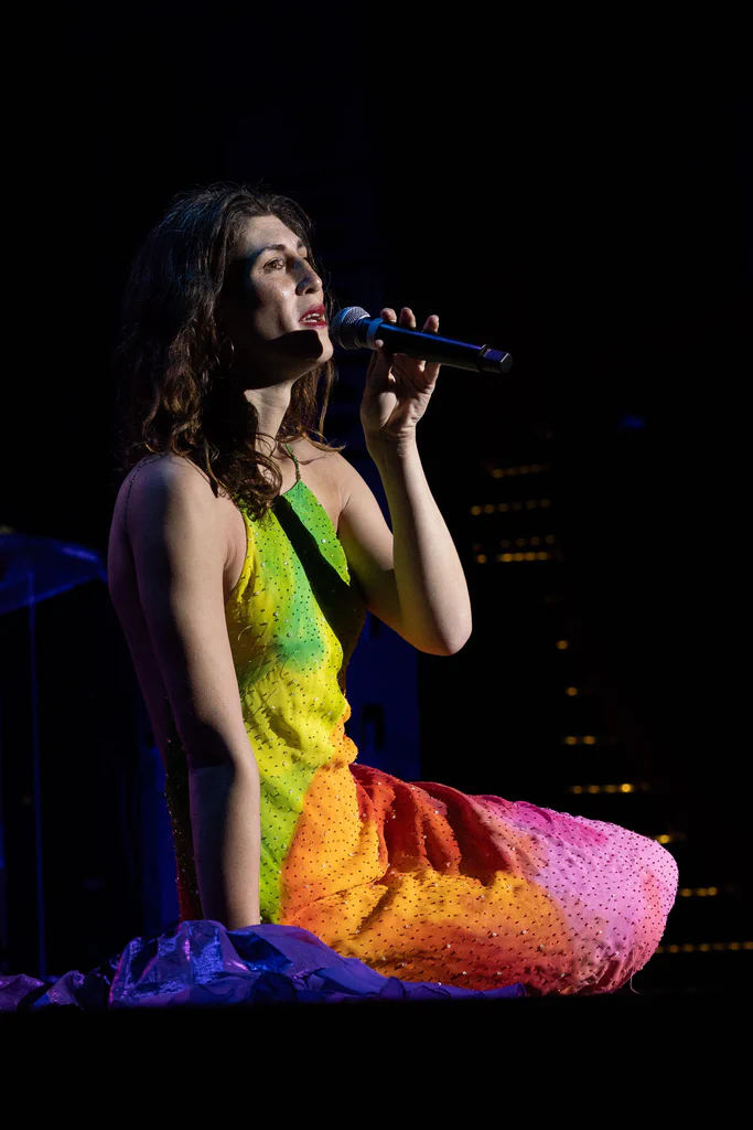 Daphne Always, a woman with wavy dark hair, is kneeling on stage in a colorful, rainbow-hued dress while singing into a microphone during JoCo Cruise 2025.