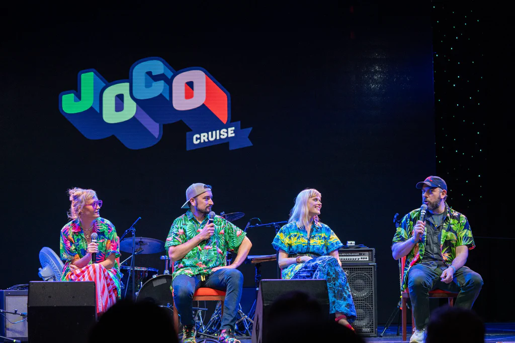 A panel of four people sitting on stage during JoCo Cruise 2025, engaging in a discussion or performance.