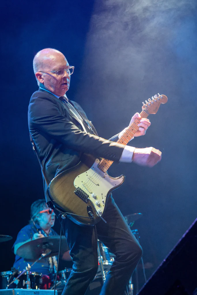 A man playing an electric guitar on stage during JoCo Cruise 2025, with a drummer in the background.