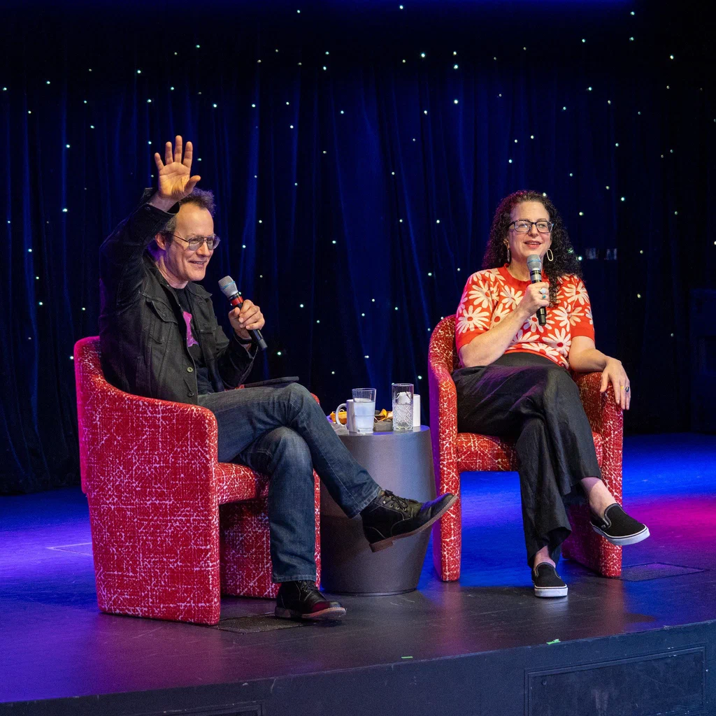 Two people seated in red chairs on stage during JoCo Cruise 2025; one is waving while holding a microphone, and the other is smiling with a microphone in hand.