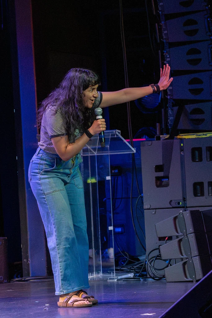 A woman on stage, holding a microphone, gesturing passionately with her right hand during a performance at JoCo Cruise 2025.