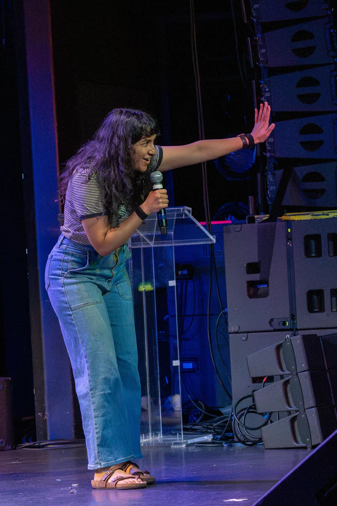 A woman on stage, holding a microphone, gesturing passionately with her right hand during a performance at JoCo Cruise 2025.