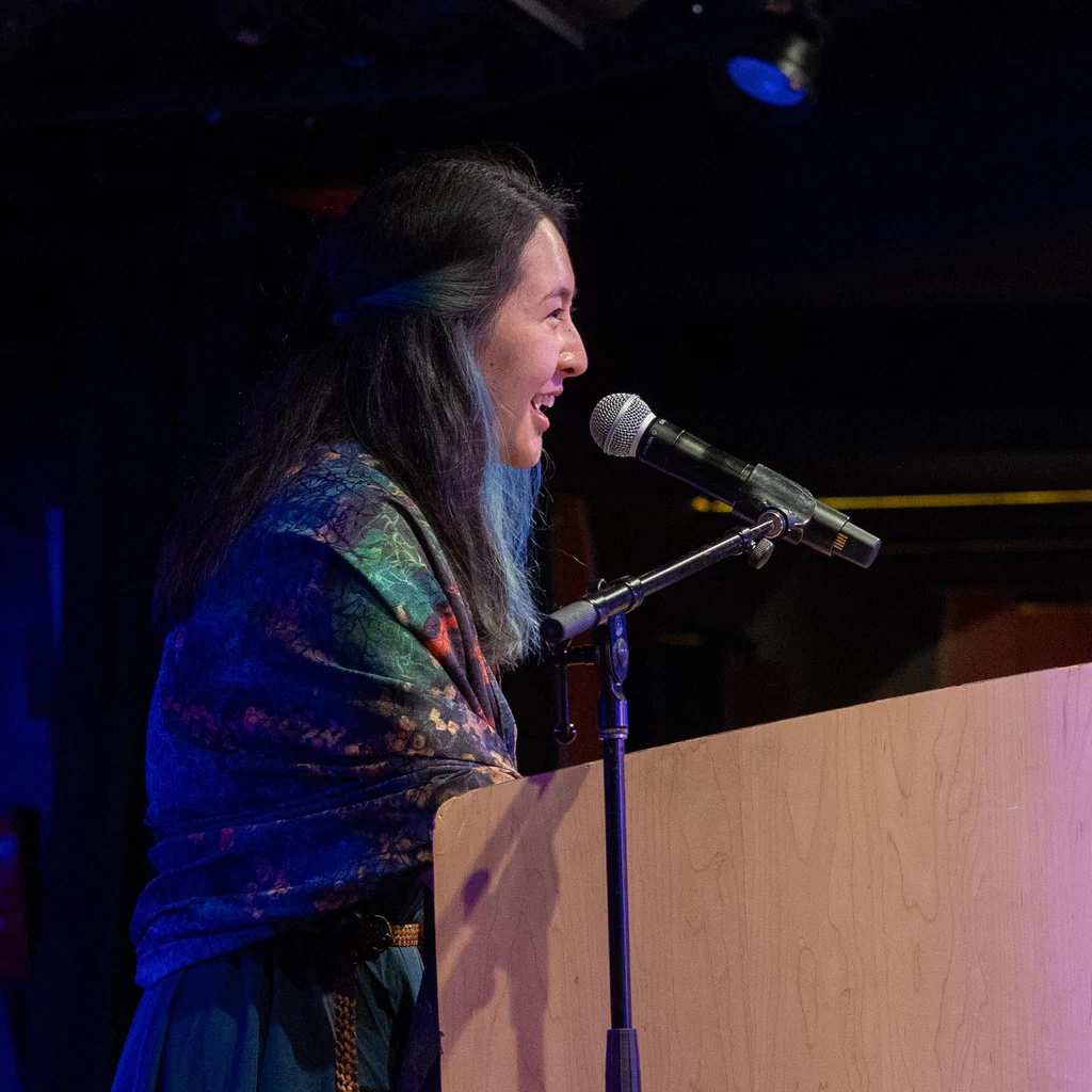 A woman with long dark hair, smiling and speaking into a microphone at a JoCo Cruise 2025 event.