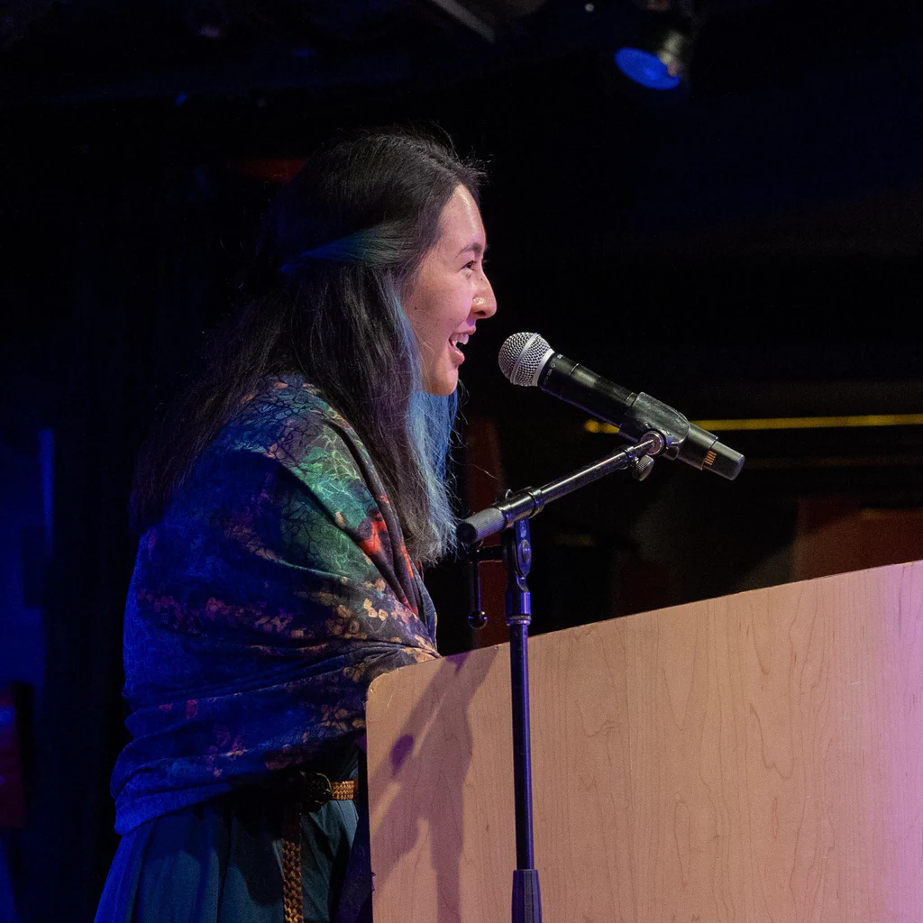 A woman with long dark hair, smiling and speaking into a microphone at a JoCo Cruise 2025 event.