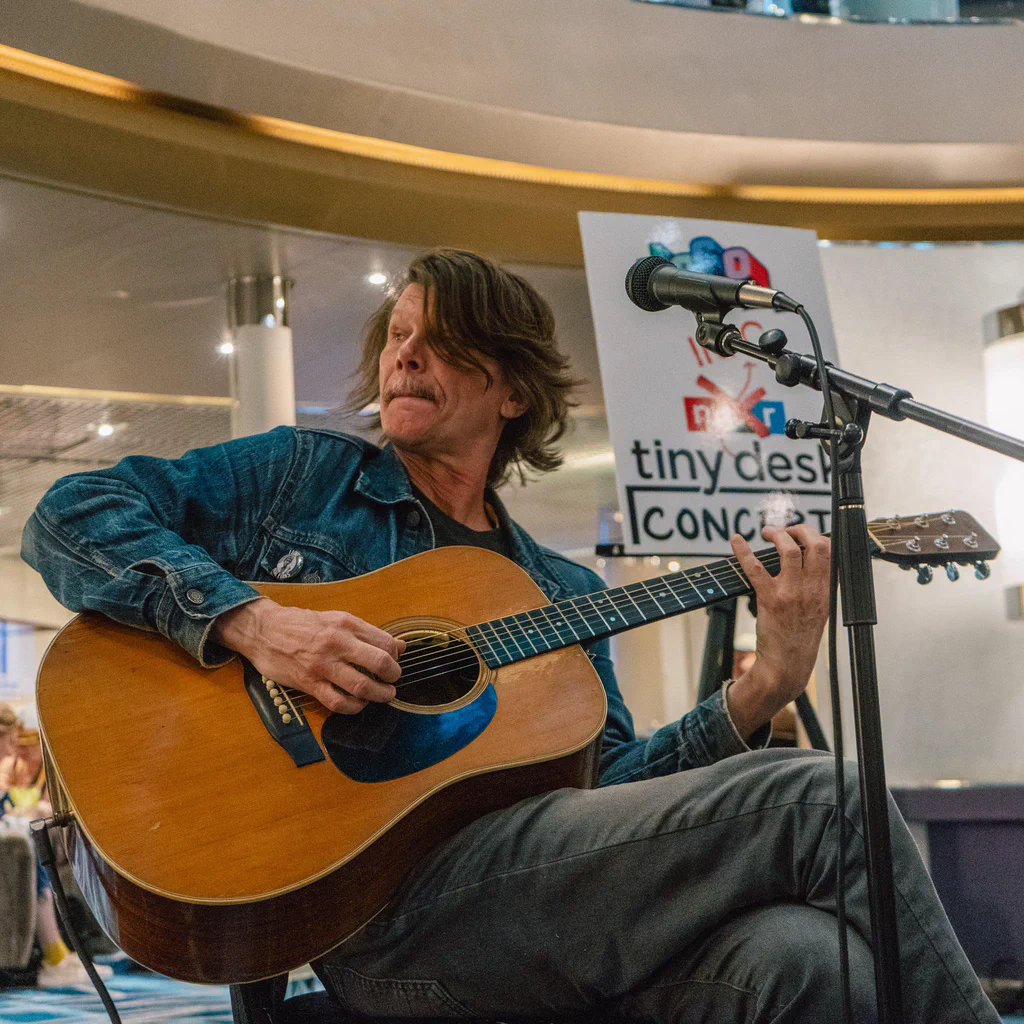 A man playing an acoustic guitar during a performance at JoCo Cruise 2025, with a "tiny desk concert" sign in the background.