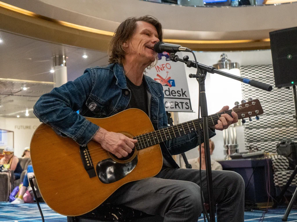 A man with shoulder-length hair playing an acoustic guitar and singing into a microphone at JoCo Cruise 2025.