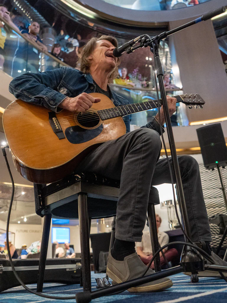 A man is sitting on a chair, playing an acoustic guitar and singing into a microphone during a performance on JoCo Cruise 2025.