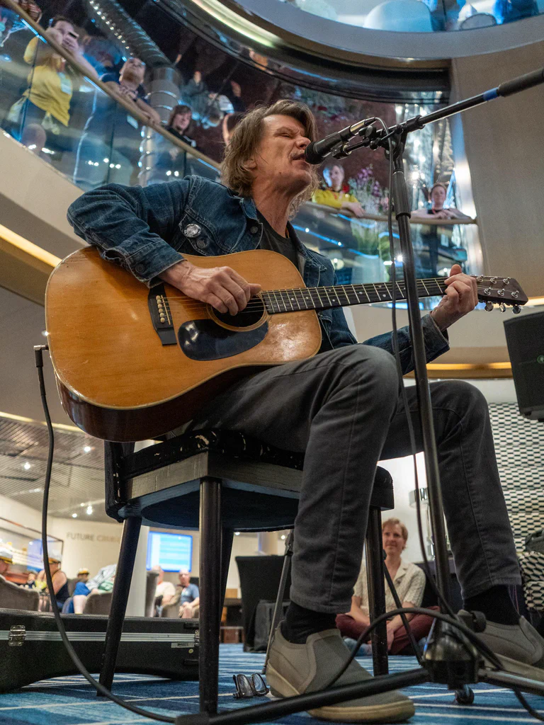 A man with shoulder-length hair wearing a denim jacket, seated on a chair, passionately singing into a microphone while playing an acoustic guitar at JoCo Cruise 2025.