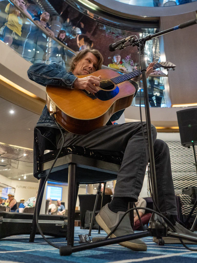 A man playing an acoustic guitar on stage at JoCo Cruise 2025, with audience members in the background.