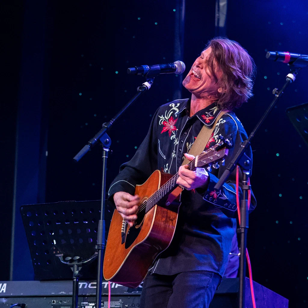 A man is performing on stage with an acoustic guitar, singing passionately into a microphone, at JoCo Cruise 2025.