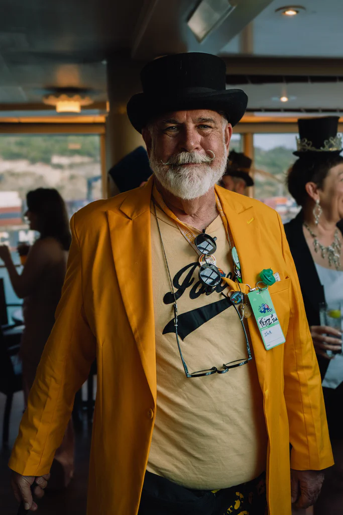 A man dressed in formal attire with a yellow jacket and a black top hat, smiling at a cruise event, with others in costume in the background.