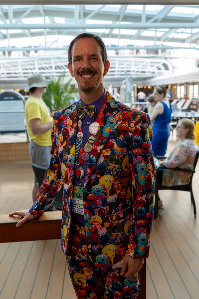 A man dressed in a colorful suit featuring Sesame Street characters stands indoors at the JoCo Cruise 2025 event, smiling at the camera.