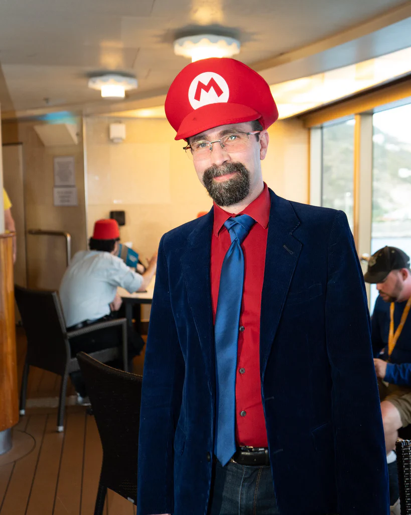 A man dressed in formal attire, wearing a red hat resembling Mario's, posing indoors at JoCo Cruise 2025.