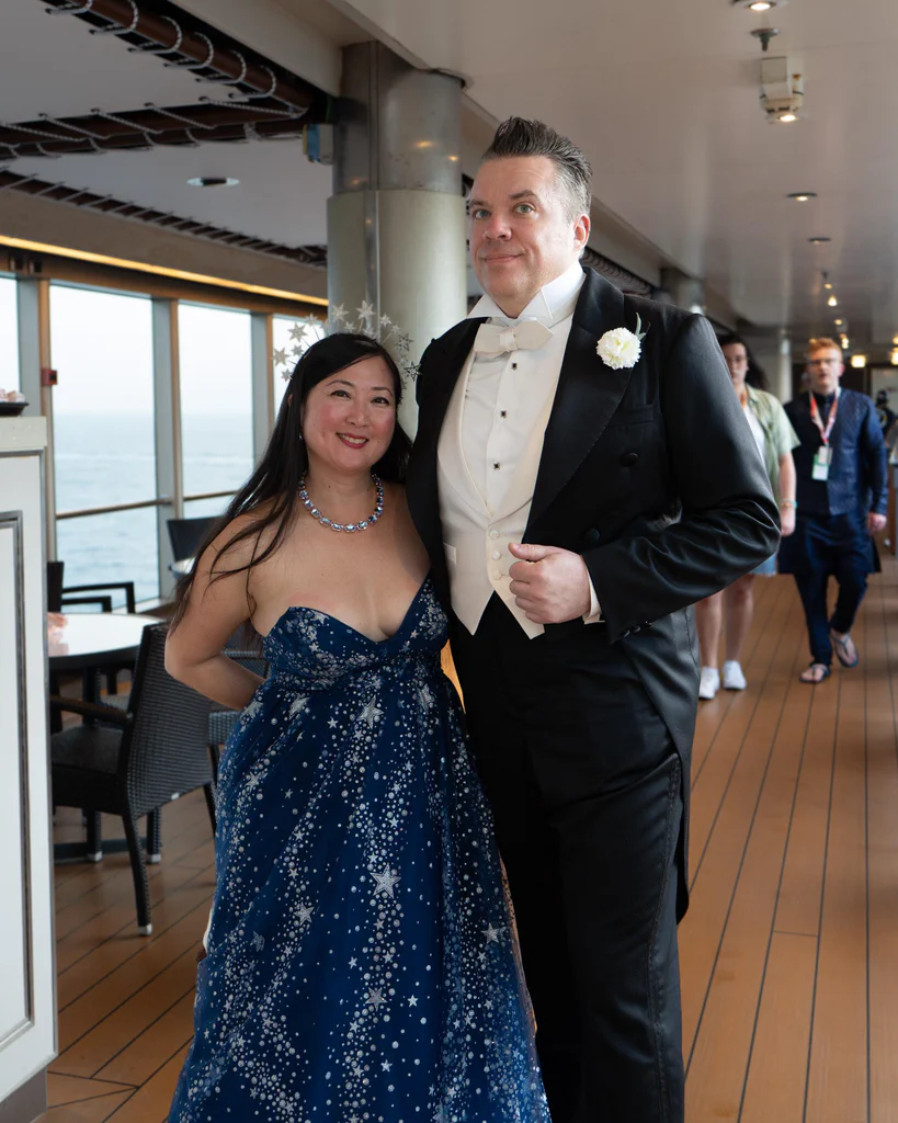 A woman in a starry blue strapless dress and a man in formal tuxedo with a white bow tie pose together on the JoCo Cruise 2025.