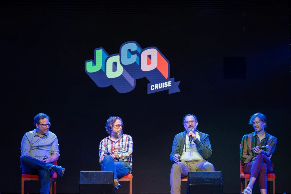 A panel of four men sitting on stage at JoCo Cruise 2025, with a "JoCo Cruise" logo projected on the screen behind them.