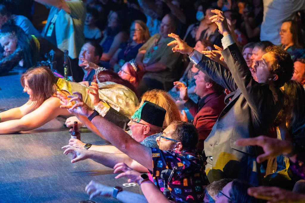 An audience at JoCo Cruise 2025, eagerly watching and reaching out towards a performance or event on stage, with some dressed in costumes and a lively atmosphere.