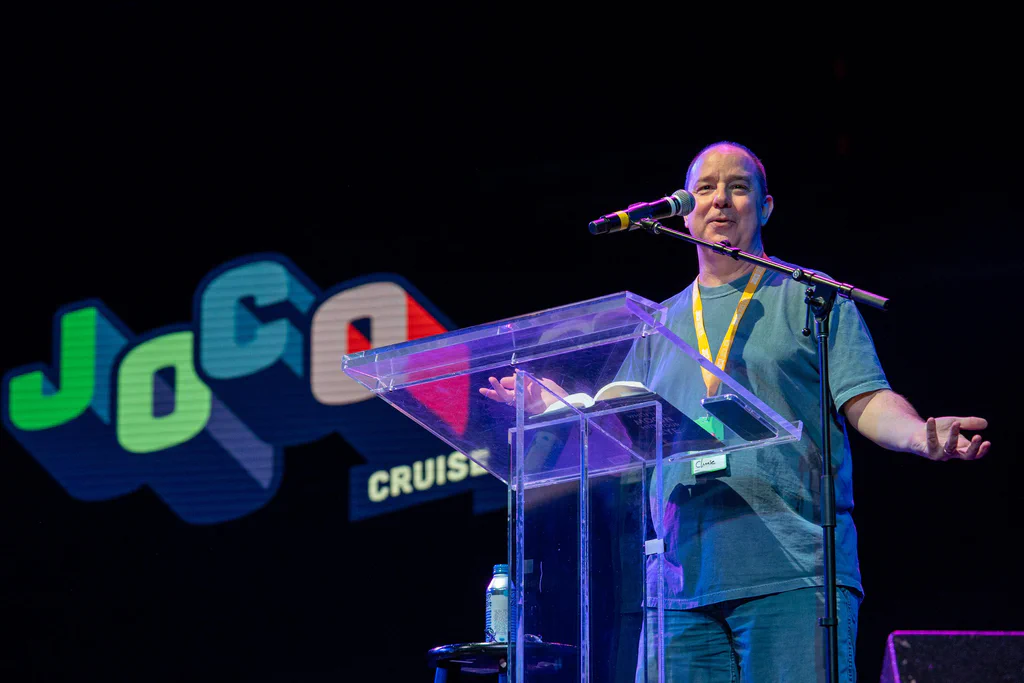 A person standing behind a clear podium on stage at JoCo Cruise 2025, speaking into a microphone.