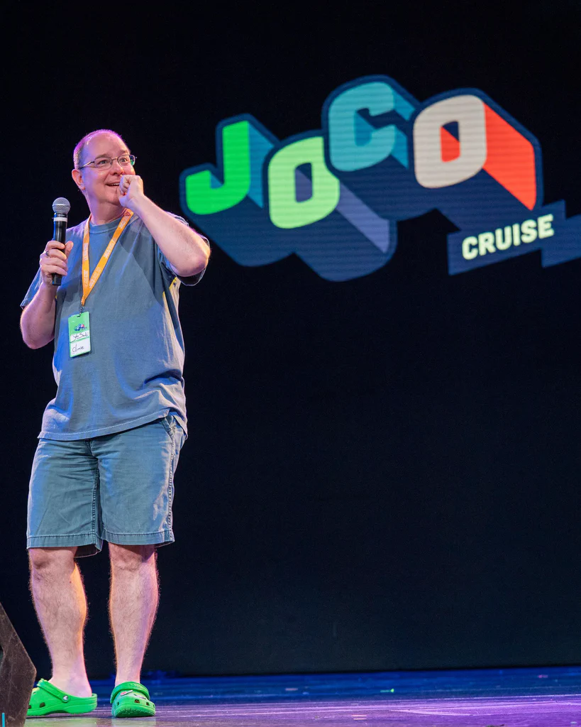 A person holding a microphone and touching their face, standing on stage with a "JoCo Cruise" logo in the background.
