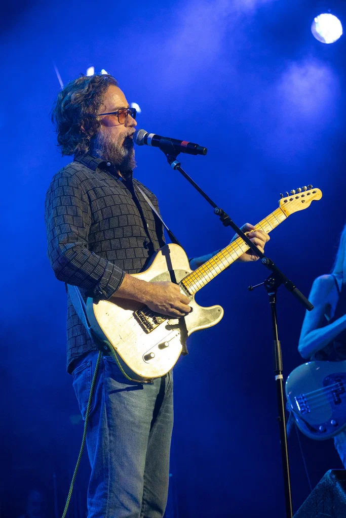 A man with curly hair and a beard, wearing glasses and a patterned shirt, playing an electric guitar on stage with blue lighting during JoCo Cruise 2025.